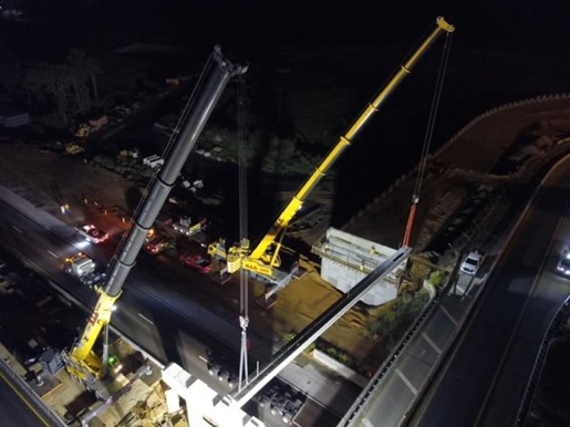 Two cranes lifting a large girder at night next to a road.